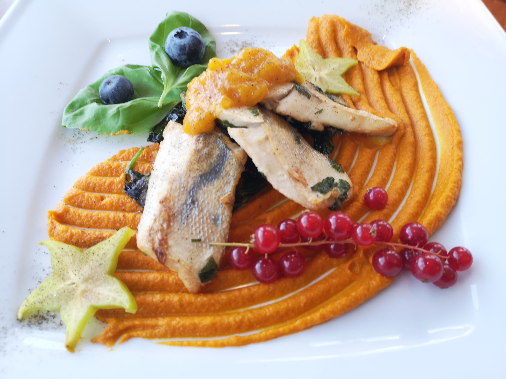 Wykwintne danie na białym talerzu: smażona ryba z pomarańczowym puree, sosem mango, karambolą, czerwoną porzeczką i borówkami na liściach bazylii.