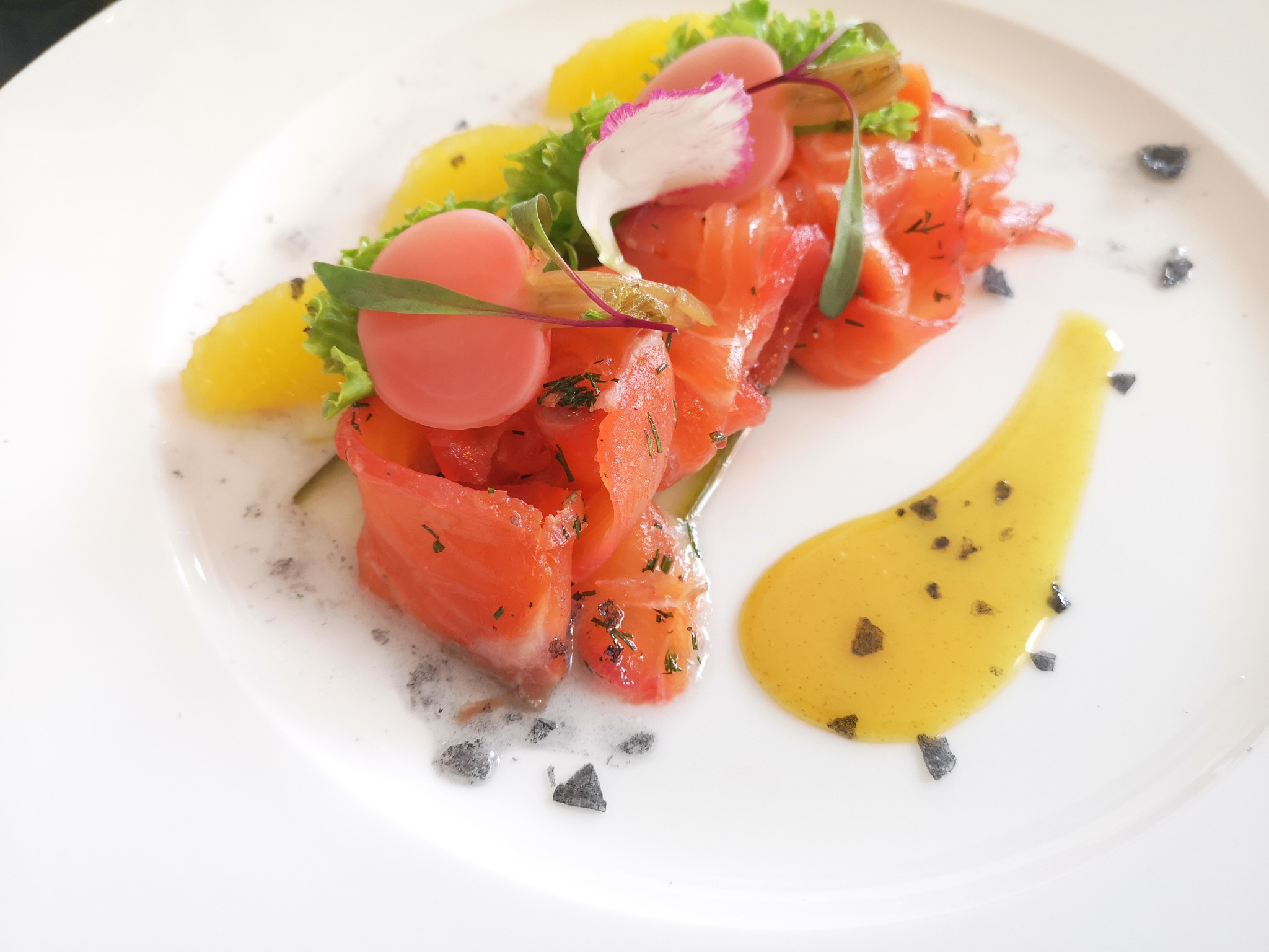 Elegancko podane danie z łososia z pomarańczami, dekorowane listkami sałaty, różowymi kulkami i sosem na białym talerzu.