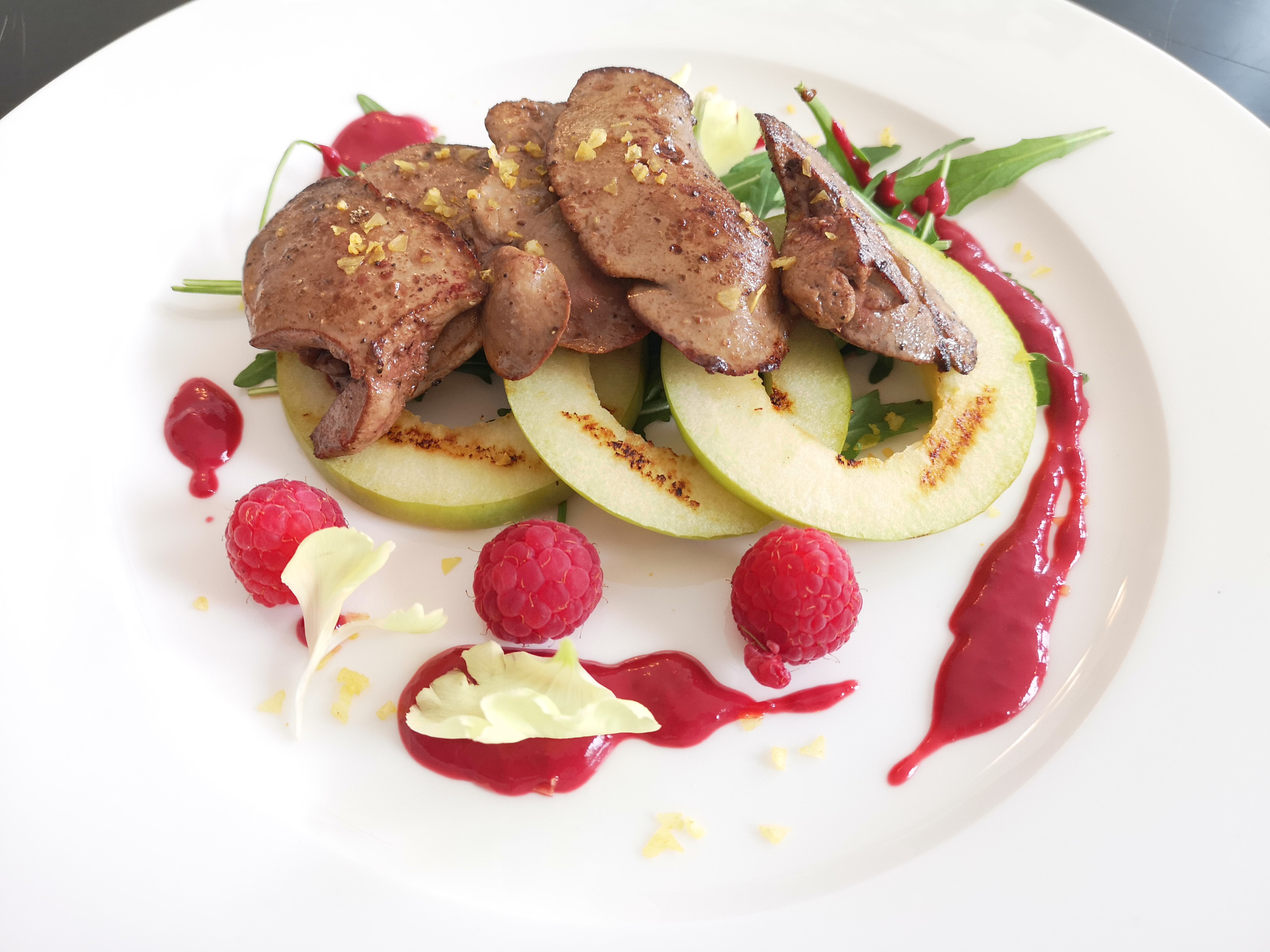 Elegancko podane danie: grillowane plastry jabłka, smażona wątróbka posypana przyprawami, maliny i sos malinowy na białym talerzu.