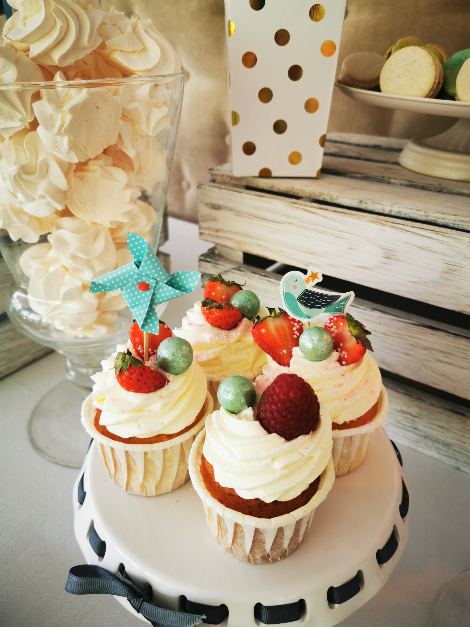 Cztery babeczki z kremem, truskawkami i maliną na białym paterze z dekoracjami, w tle słoik z bezami i pudełko w złote kropki