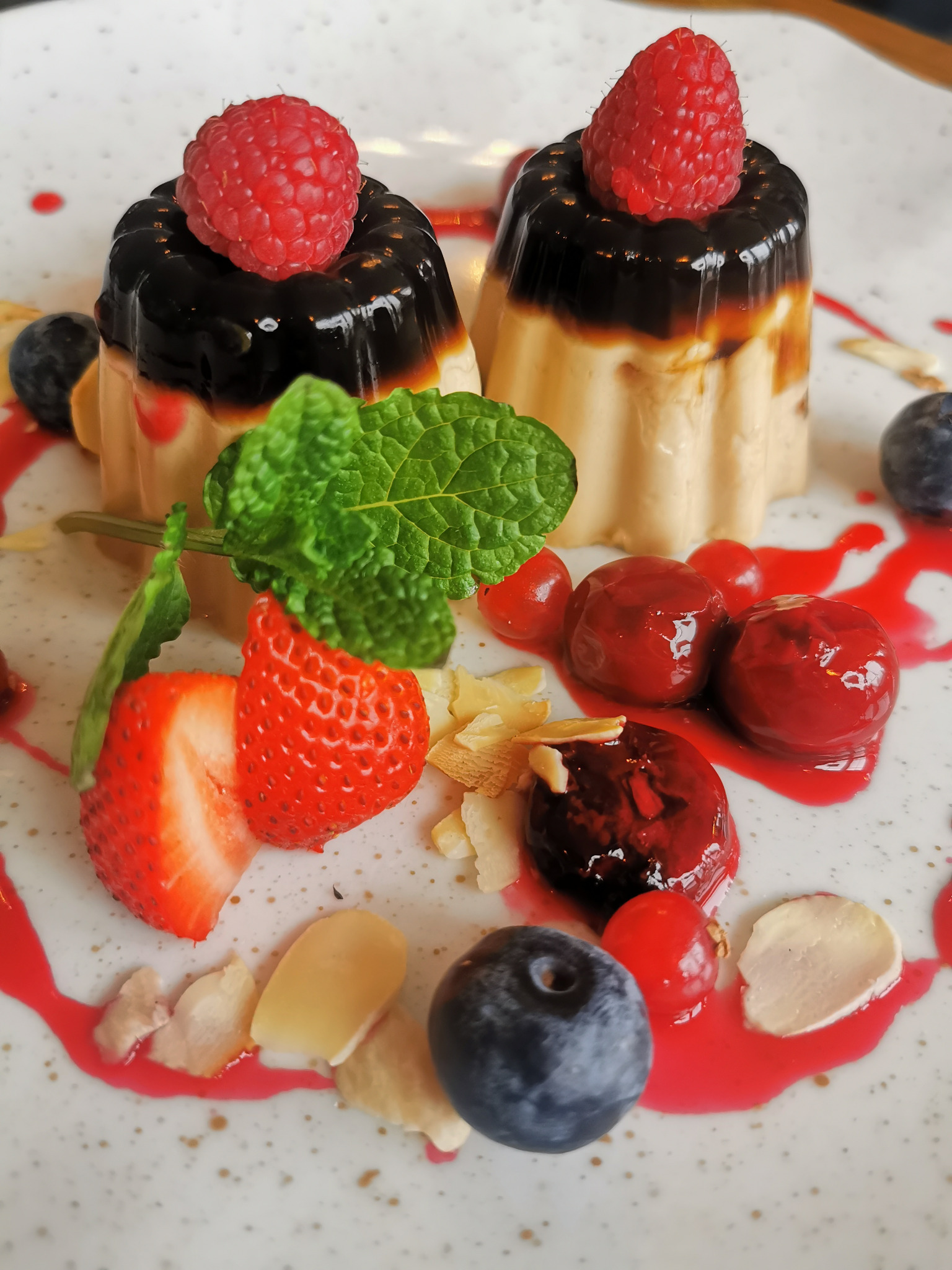 Dwa desery panna cotta z czarną polewą i malinami na wierzchu, otoczone świeżymi owocami: truskawkami, borówkami, wiśniami, czerwoną porzeczką, listkami mięty oraz płatkami migdałów, polane...