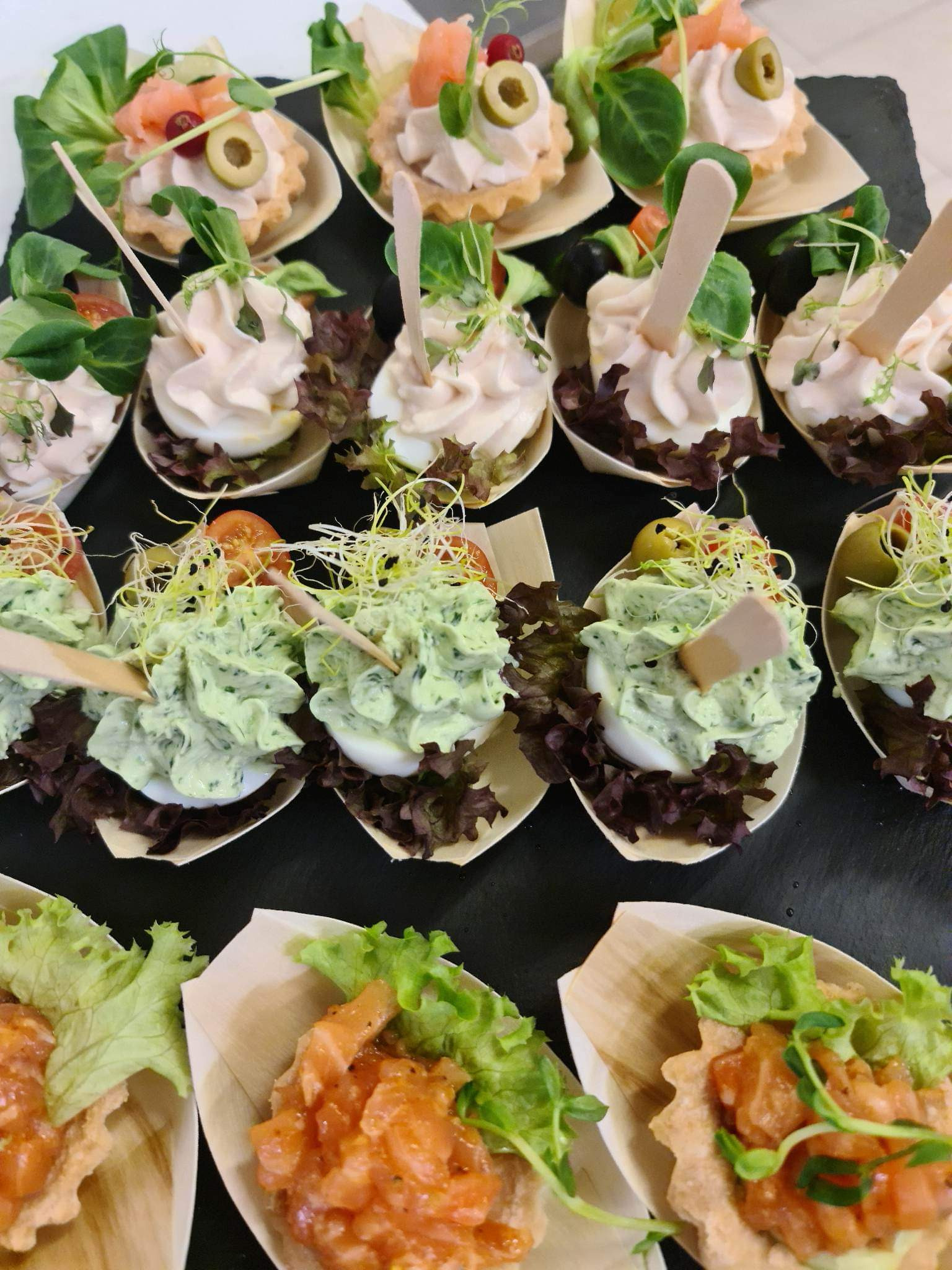 Eleganckie przekąski: jajka z różnymi pastami i dodatkami, podane na drewnianych łyżeczkach na czarnym tle.