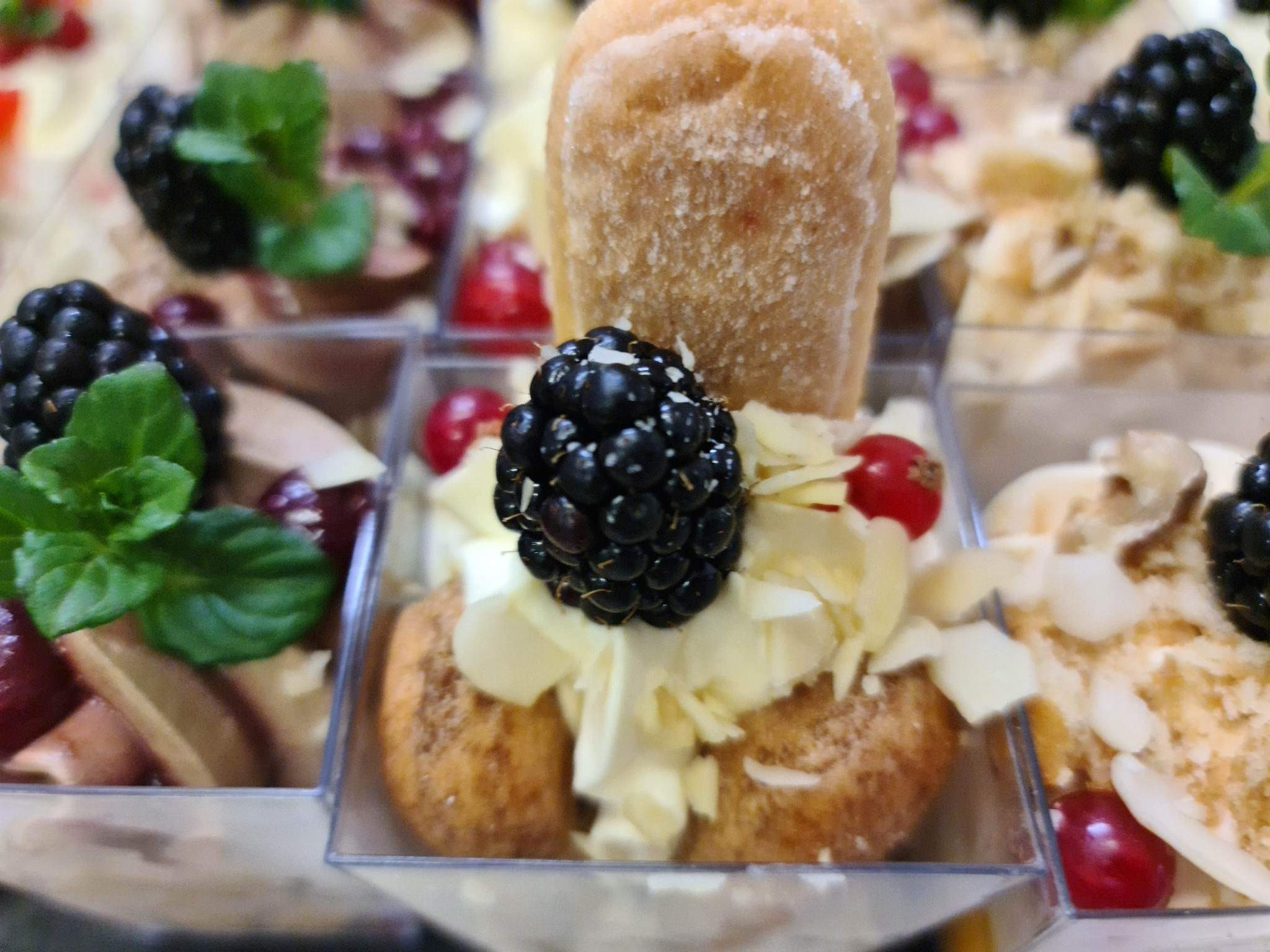 Mini deserki w przezroczystych pojemniczkach: biszkopt, krem, płatki migdałów, jeżyna i czerwona porzeczka, widoczne inne desery w tle.