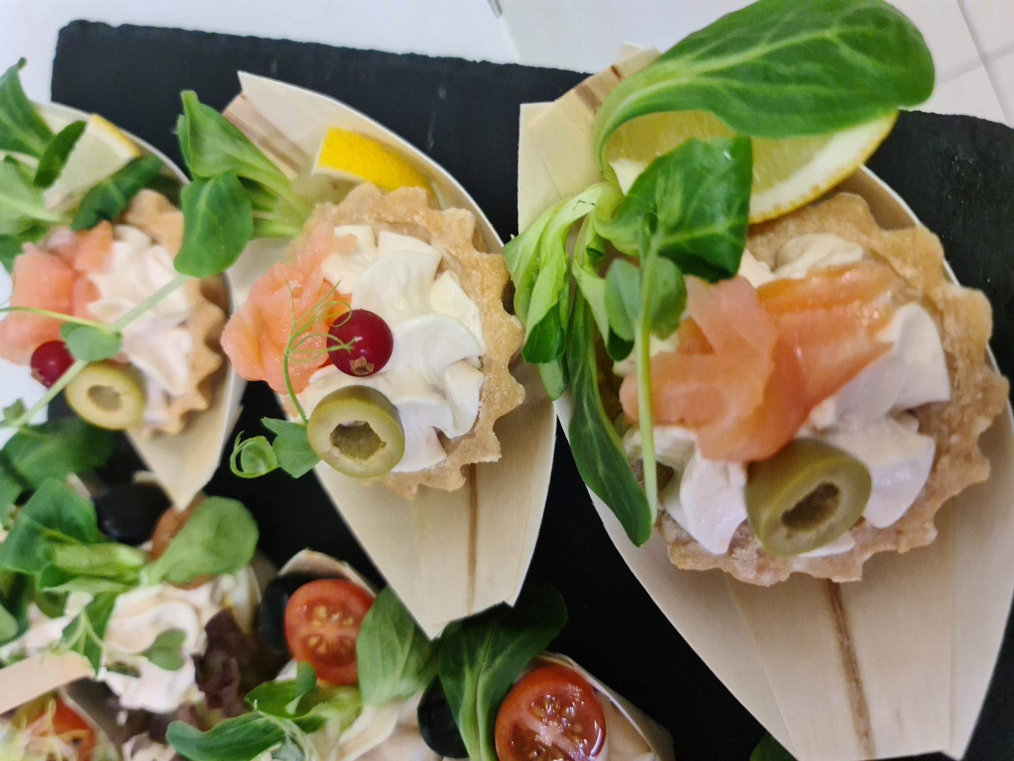 Eleganckie tartaletki z kremowym serem, łososiem, oliwkami, czerwoną porzeczką i listkami sałaty, podane na czarnym, matowym tle