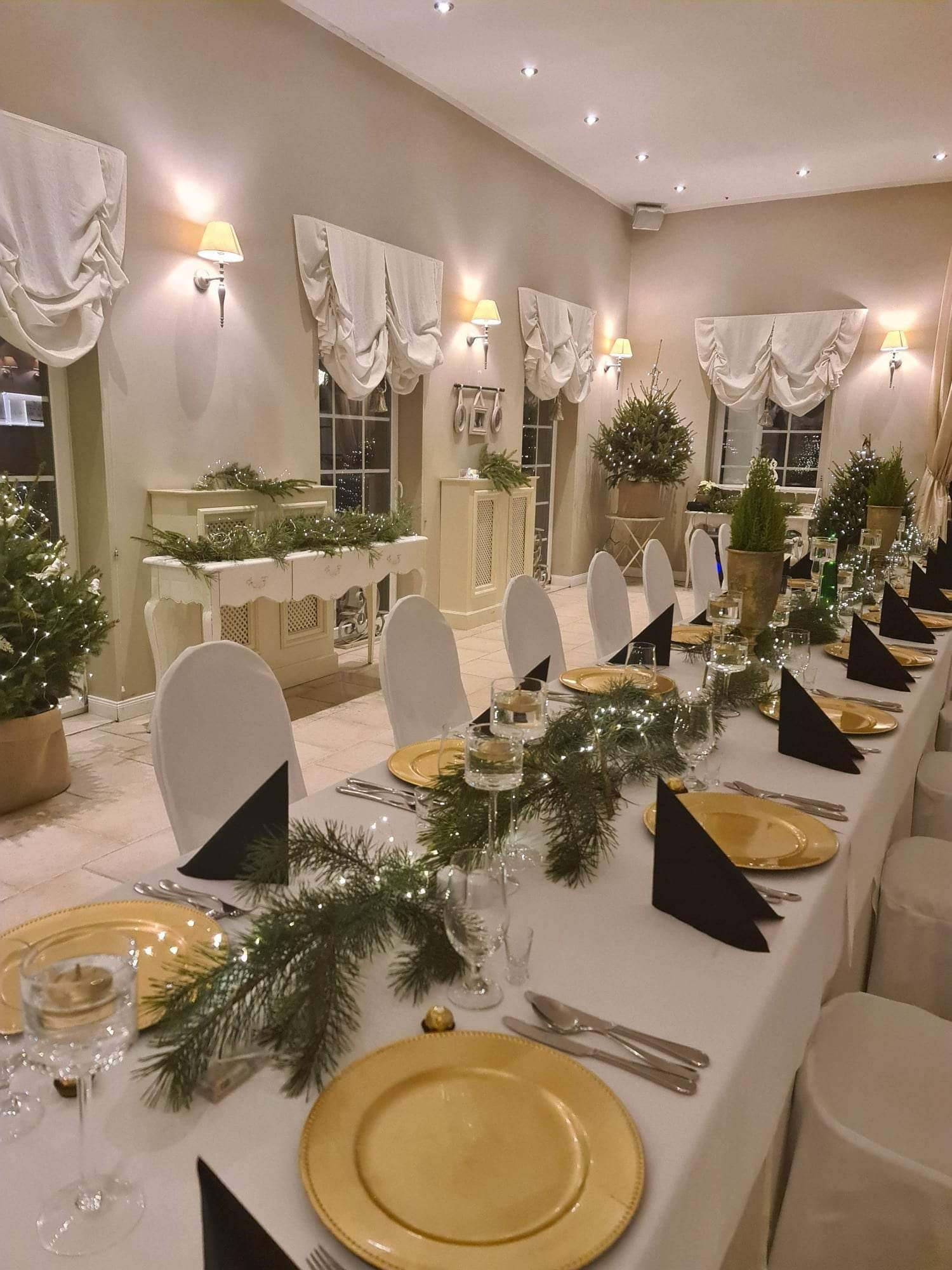 Elegancko nakryty stół na przyjęcie świąteczne z białym obrusem, złotymi talerzami, czarnymi serwetkami, gałązkami świerkowymi z lampkami i choinkami w tle.