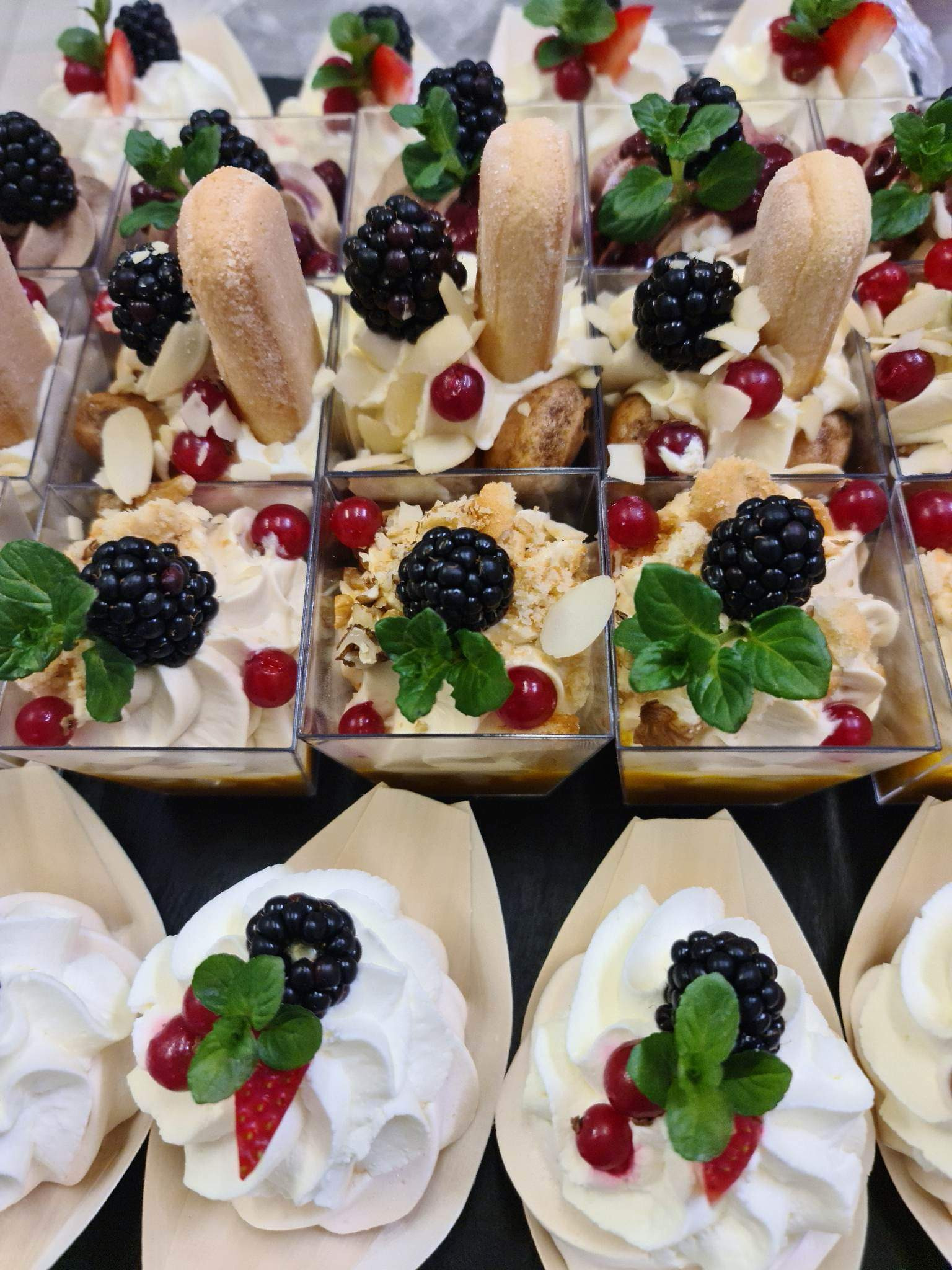 Różnorodny wybór mini deserów: kremowe musy w przezroczystych pojemnikach, jedne z biszkoptami i migdałami, inne w drewnianych łódeczkach, wszystkie udekorowane jeżynami, czerwoną porzeczką...