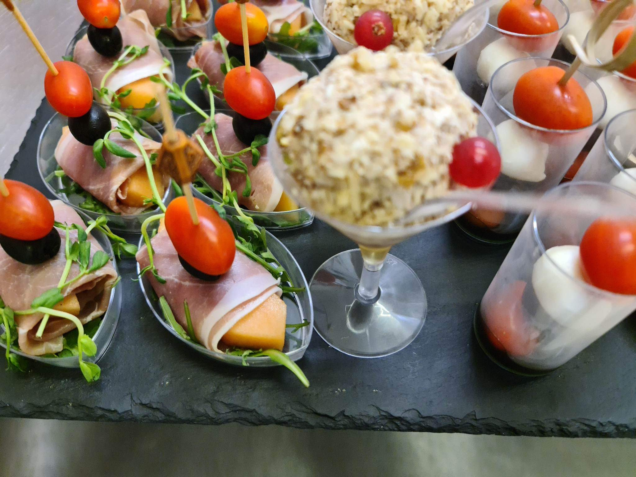 Elegancko podane przekąski: melon owinięty szynką parmeńską z pomidorkiem i oliwką na wykałaczce, sałatka w kieliszku i mozzarella z pomidorkiem w szklance, ułożone na czarnym, kamiennym talerzu.