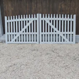 Dwie części jasnoszarej, drewnianej bramy ogrodowej z pionowymi szczebelkami i ukośnymi wzmocnieniami, leżące na żwirze na tle ciemnobrązowej, drewnianej ściany.