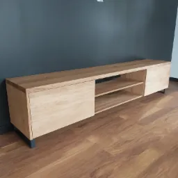 Drewniana szafka RTV z jasnego drewna, z przesuwanymi drzwiczkami i otwartymi półkami, na czarnych nóżkach, wykonana na wymiar w Sobkowie; na tle szara ściana i panele podłogowe.