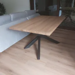 Stół z blatem z jasnego drewna i czarną, metalową podstawą, ustawiony w salonie obok szarej sofy i okna z żaluzjami.