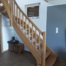 Jasne, drewniane schody z zakrętem, balustrada z pionowych szczebelków, widoczne oświetlenie w ścianie, na dole skrzynia z dekoracjami.