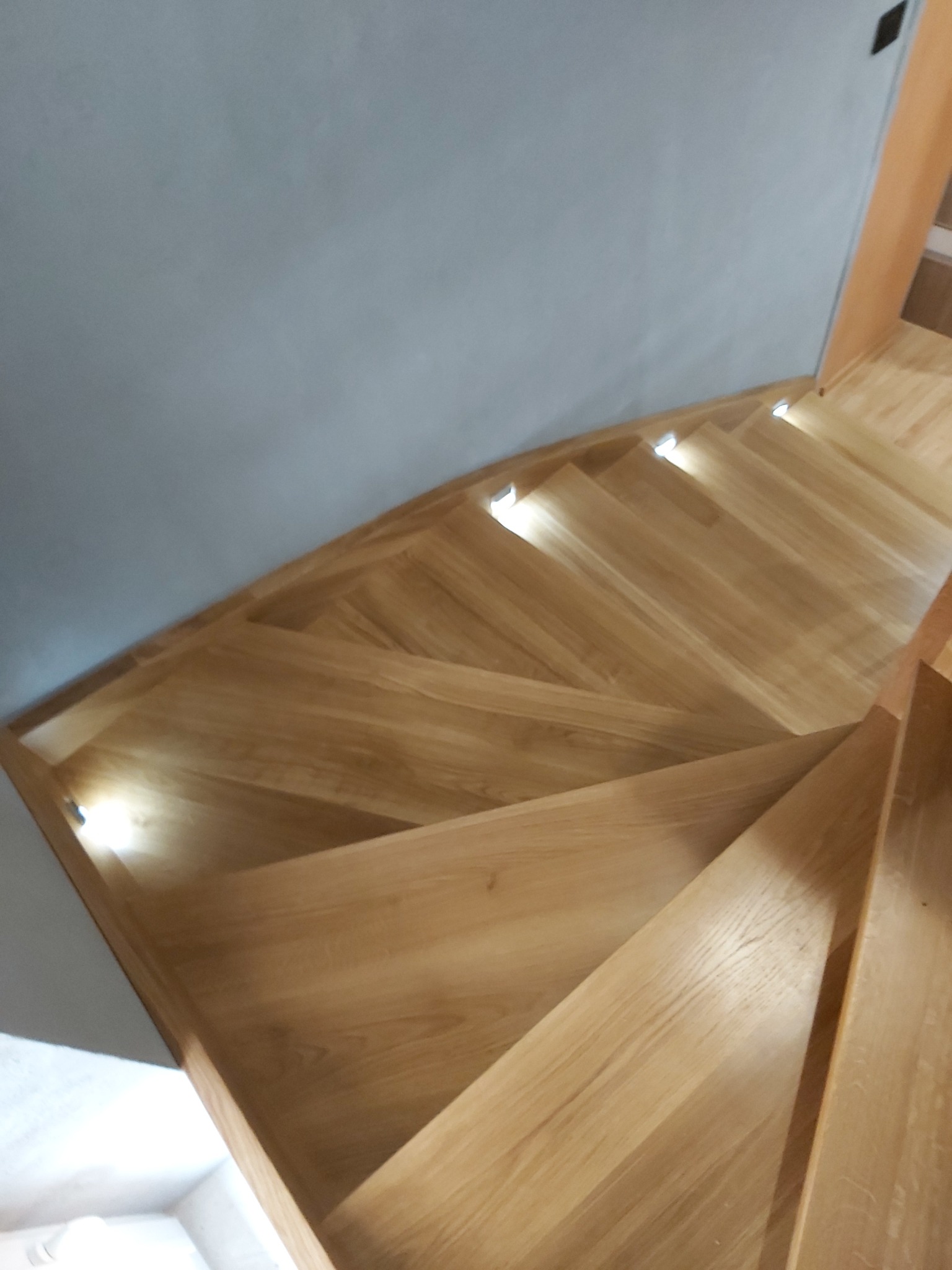 Zakręcone schody drewniane z podświetleniem LED w stopniach, widok z góry, szara ściana w tle.