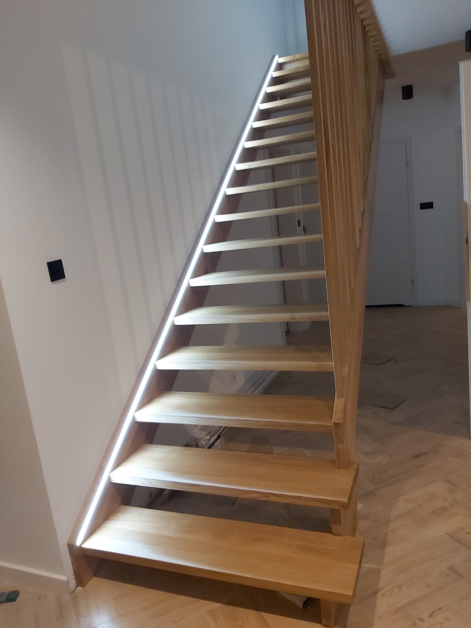 Jasne, drewniane schody z podświetleniem LED wzdłuż boku, prowadzące do góry w nowoczesnym wnętrzu z parkietem w jodełkę.