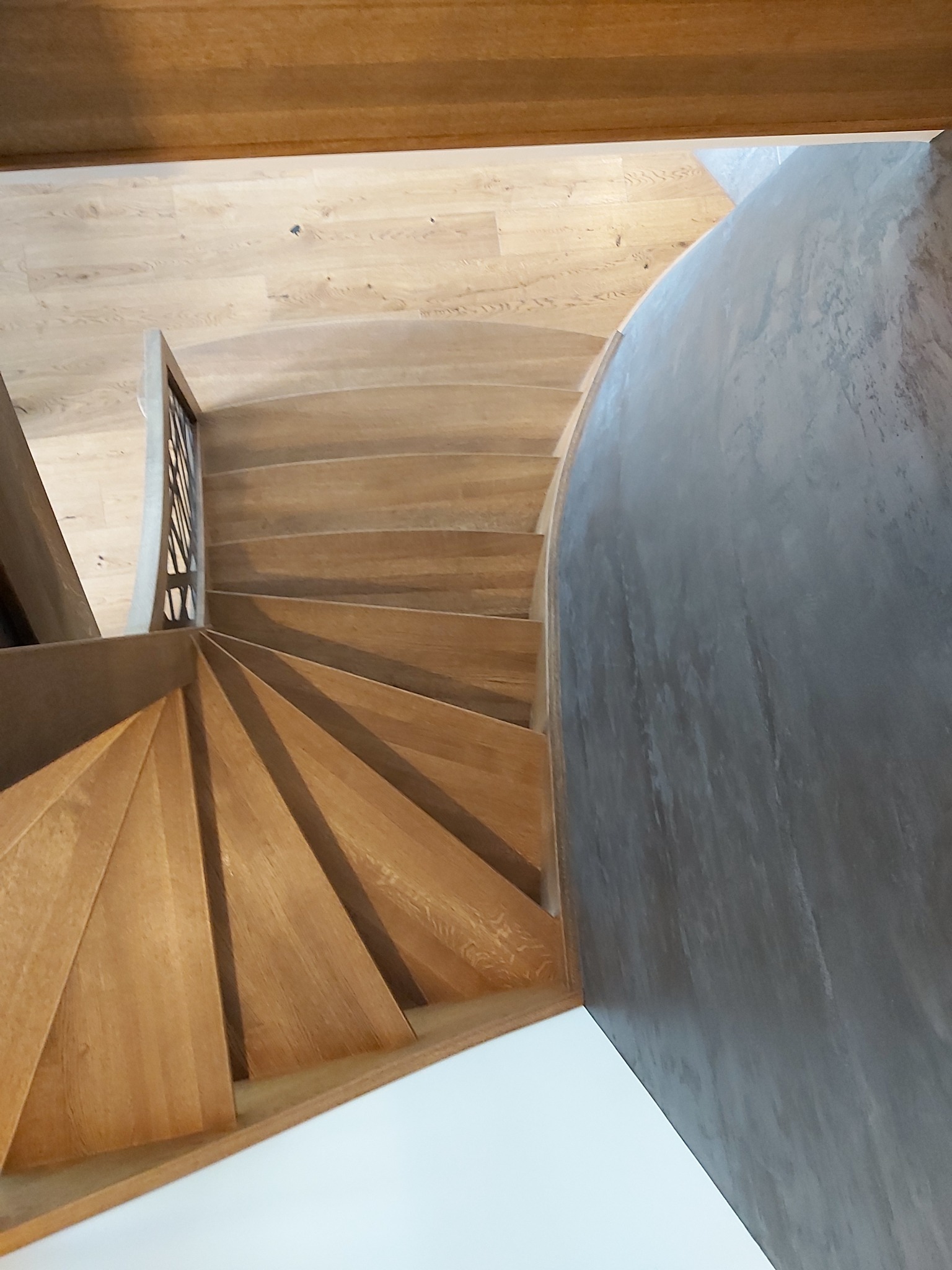 Widok z góry na kręte, drewniane schody z jasnego drewna, z elementami balustrady i szarą ścianą o fakturze betonu.