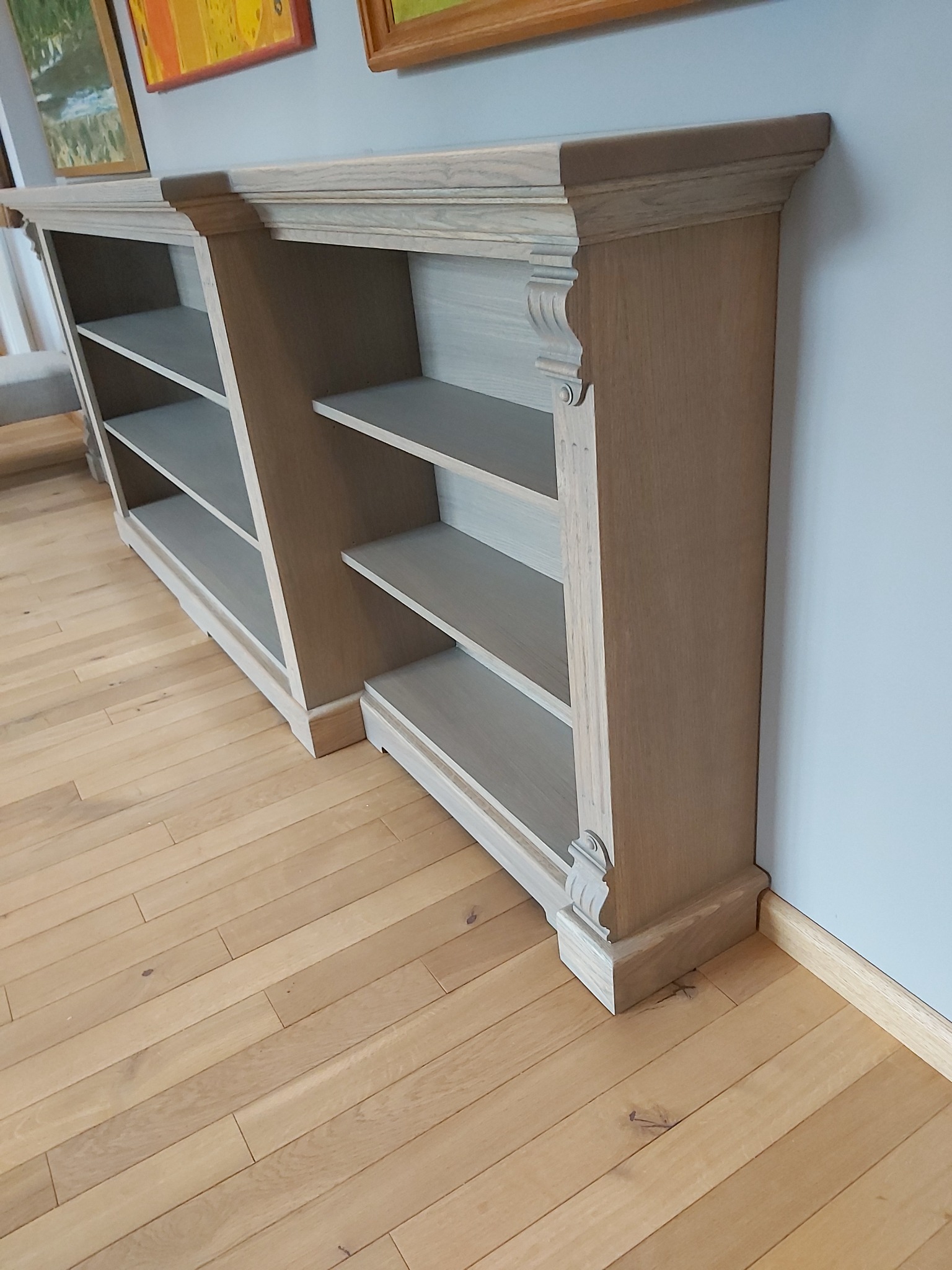 Dwie niskie, drewniane biblioteczki z półkami, w stylu klasycznym, z dekoracyjnymi elementami wykończeniowymi, ustawione na parkiecie w jasnym odcieniu drewna, z obrazami w tle.