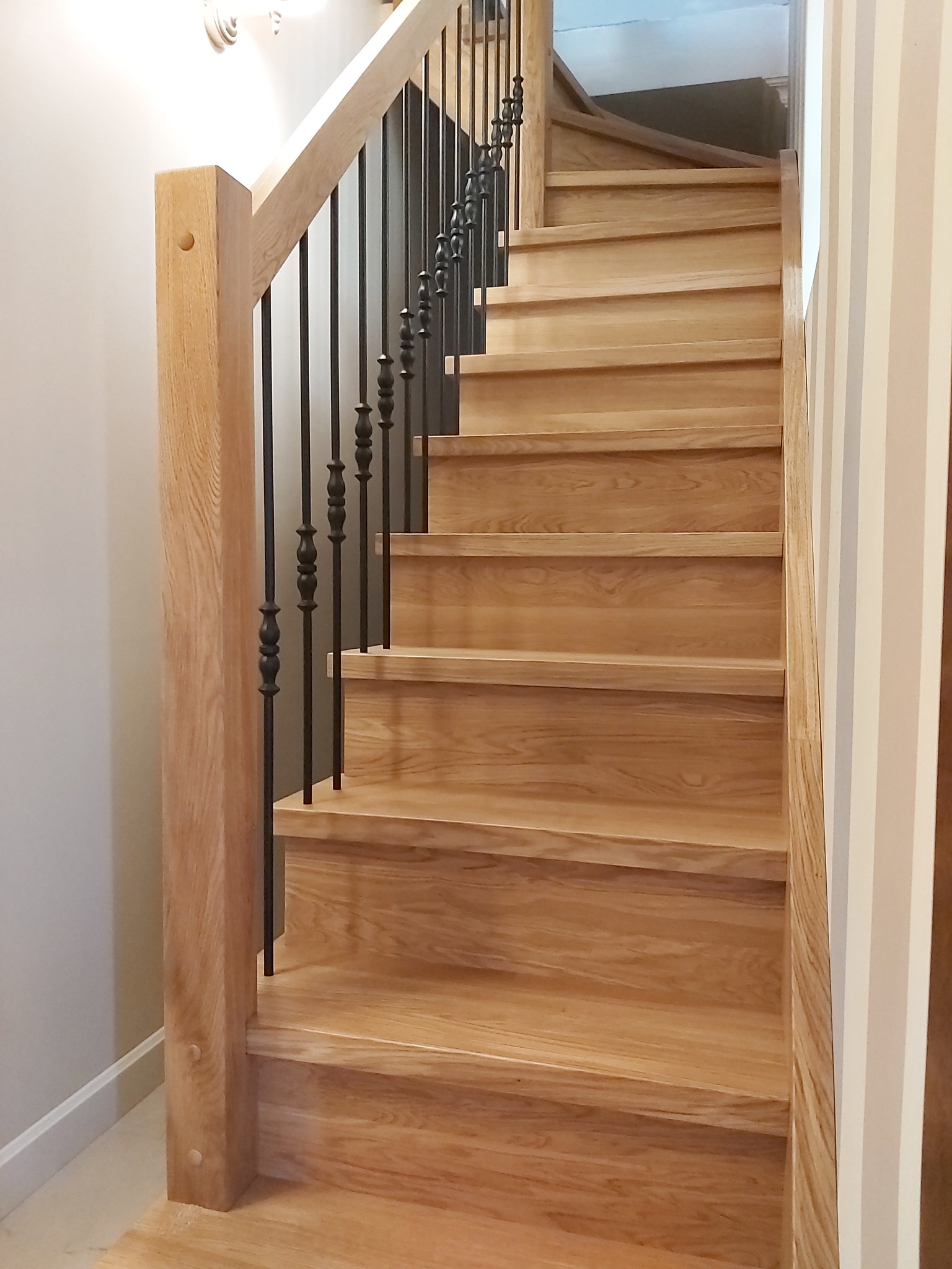 Jasne, drewniane schody z czarną, metalową balustradą, widok od dołu, z fragmentem ściany w paski po prawej stronie.
