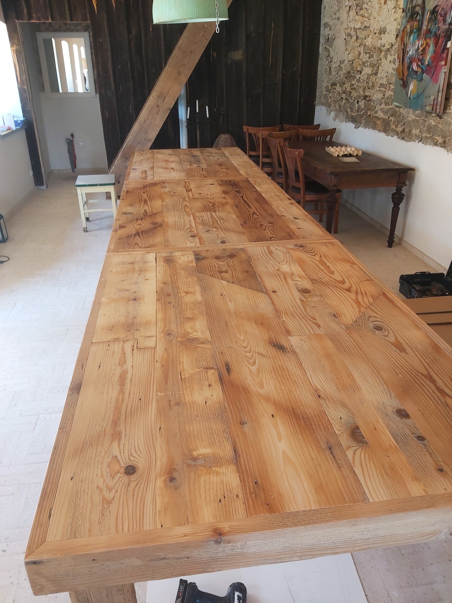 Blat stołu z jasnego drewna o widocznej strukturze słojów, składający się z kilku połączonych desek, w tle fragment wnętrza z drewnianymi elementami konstrukcyjnymi i ścianą z cegły.