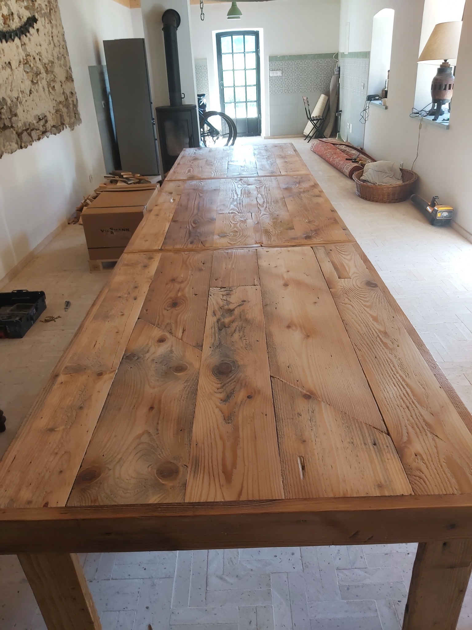 Długi, rozkładany stół z surowych desek z widocznymi słojami drewna, w jasnym wnętrzu z piecem i rowerem w tle. Stół w trakcie montażu, obok skrzynka z narzędziami i karton.