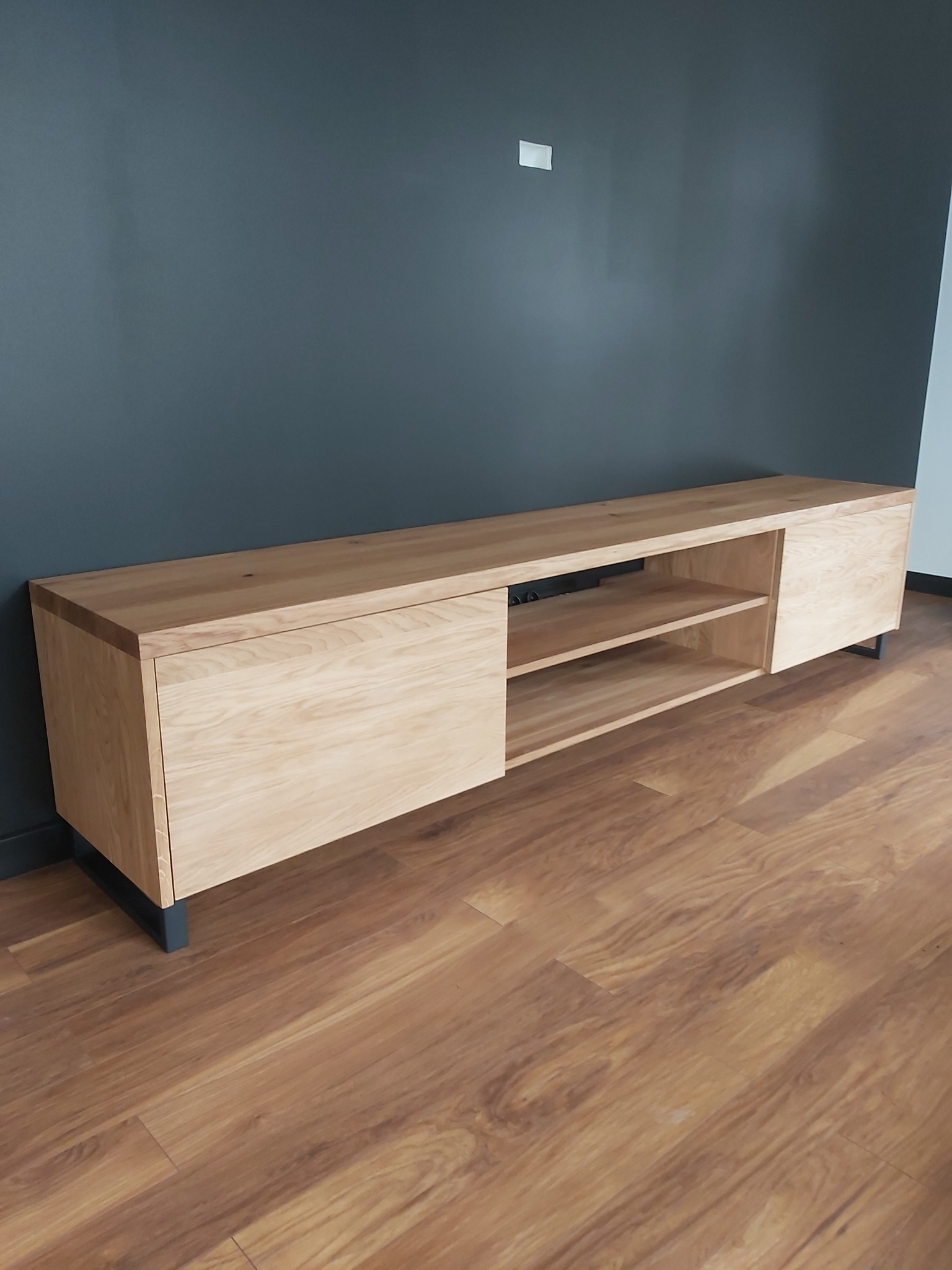 Drewniana szafka RTV z jasnego drewna, z przesuwanymi drzwiczkami i otwartymi półkami, na czarnych nóżkach, wykonana na wymiar w Sobkowie; na tle szara ściana i panele podłogowe.