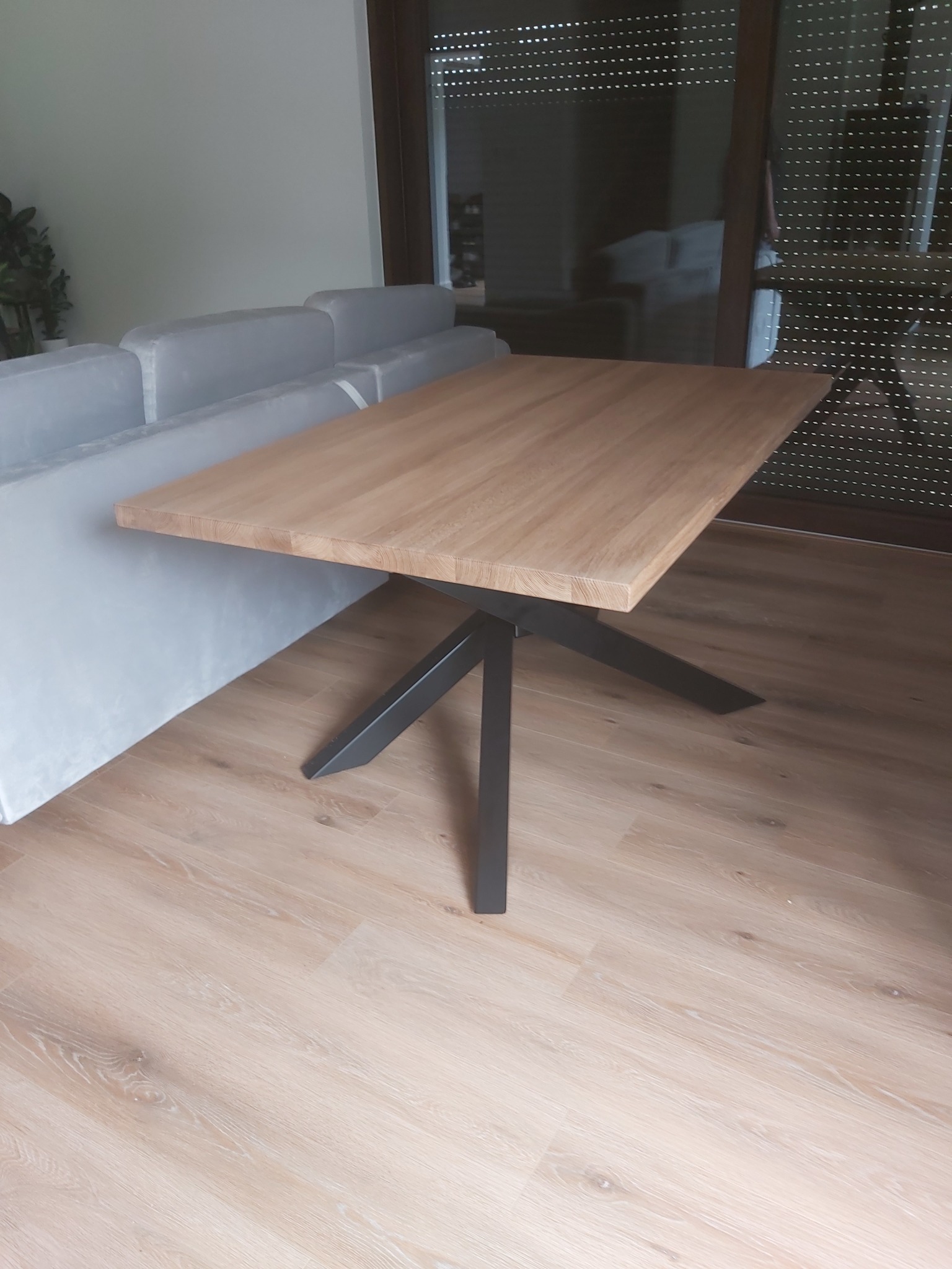 Stół z blatem z jasnego drewna i czarną, metalową podstawą, ustawiony w salonie obok szarej sofy i okna z żaluzjami.