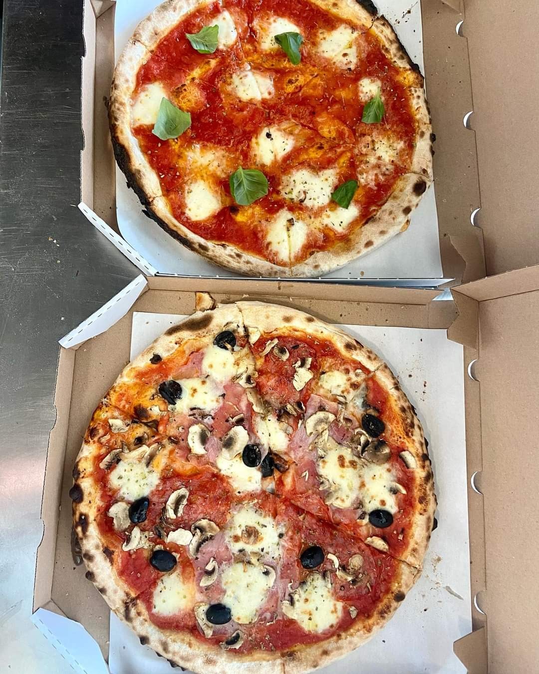 Dwie pizze w kartonowych pudełkach: jedna Margherita z bazylią, druga z szynką, pieczarkami i czarnymi oliwkami.