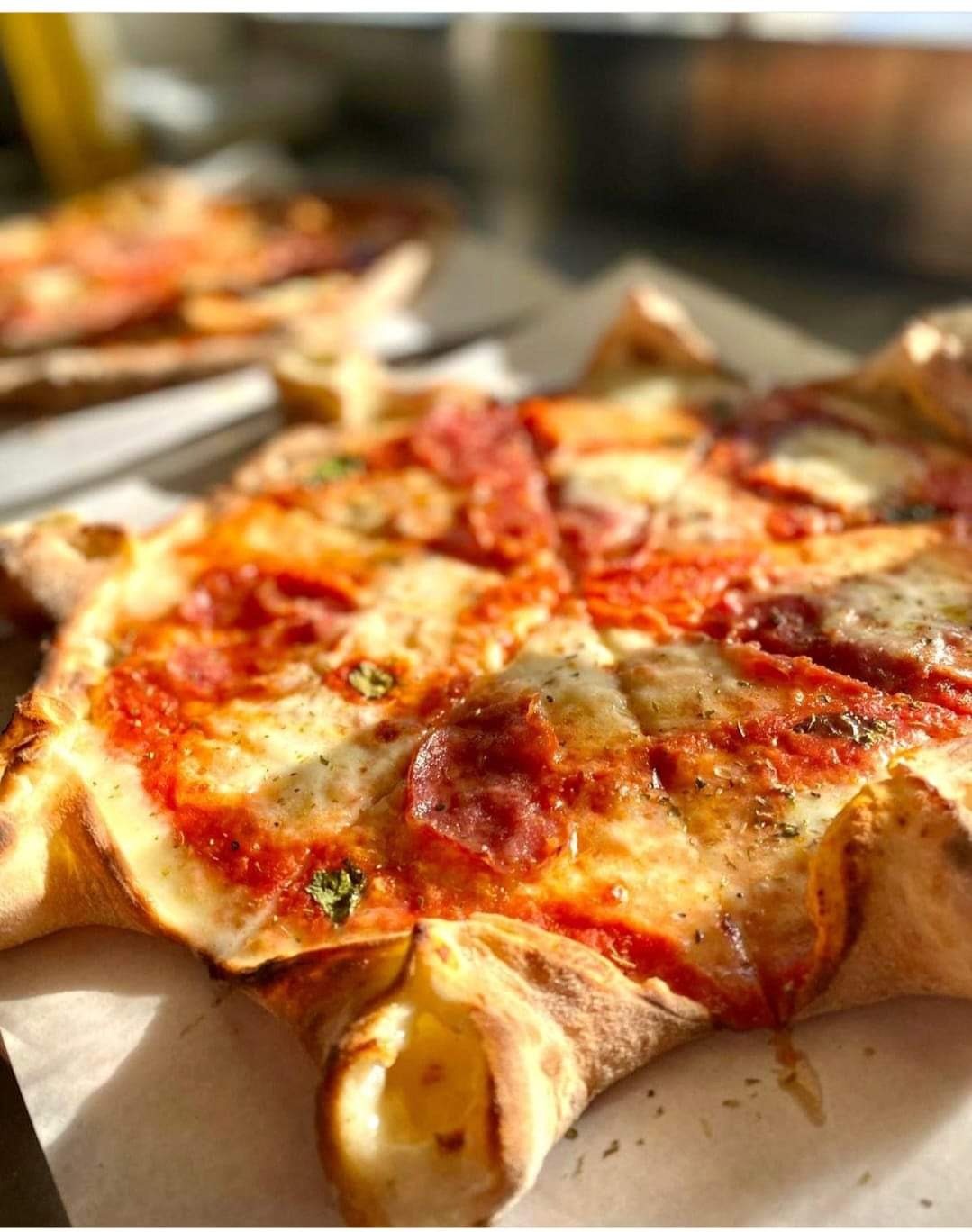 Pizza w kształcie gwiazdy z salami, serem mozzarella i sosem pomidorowym, posypana ziołami, na papierze do pieczenia, z drugą pizzą w tle, oświetlona naturalnym światłem.