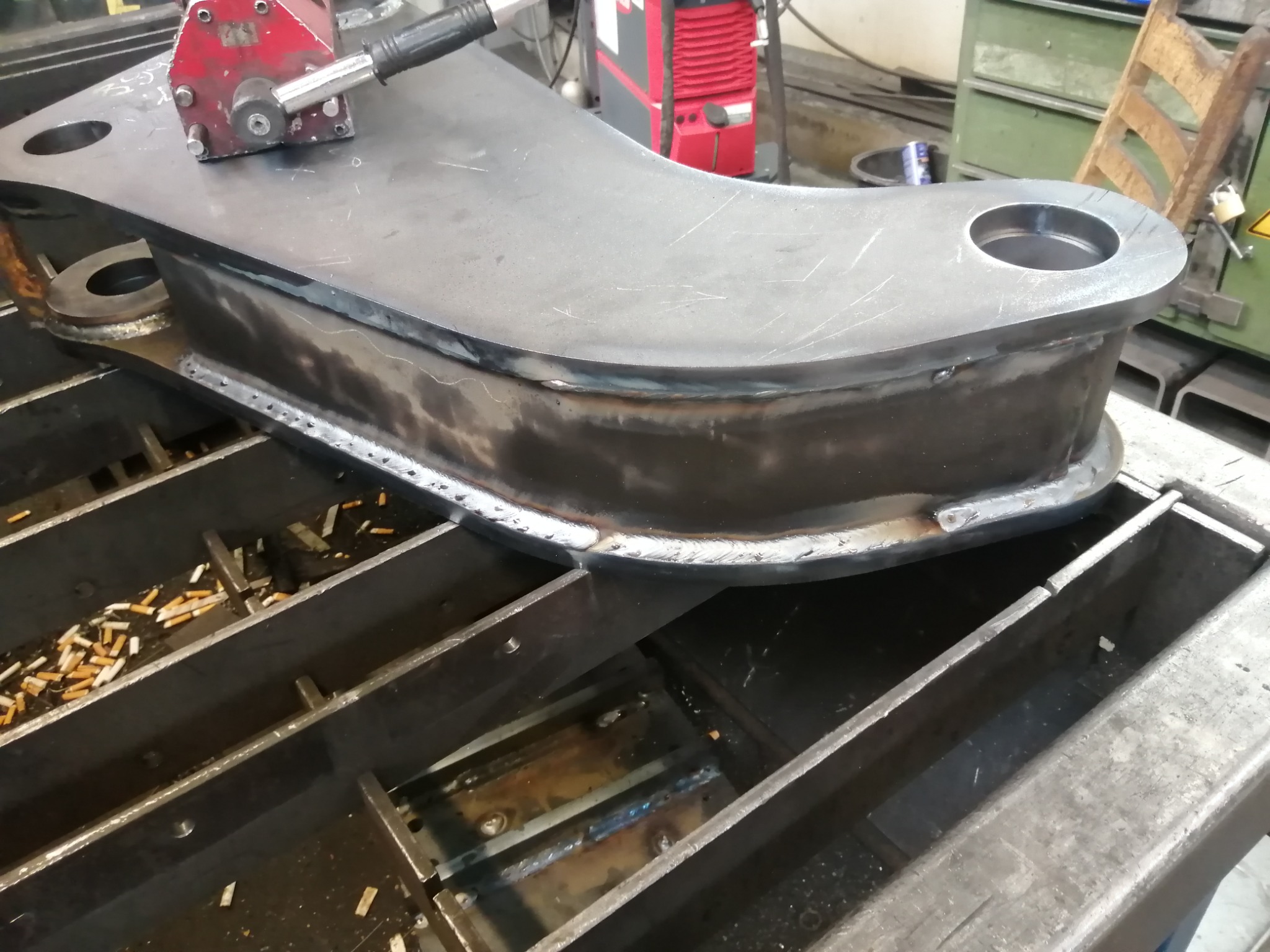 Częściowo obrobiony, spawany element konstrukcyjny z metalu, leżący na stole spawalniczym, z widocznymi śladami spawania i licznymi niedopałkami papierosów w tle.