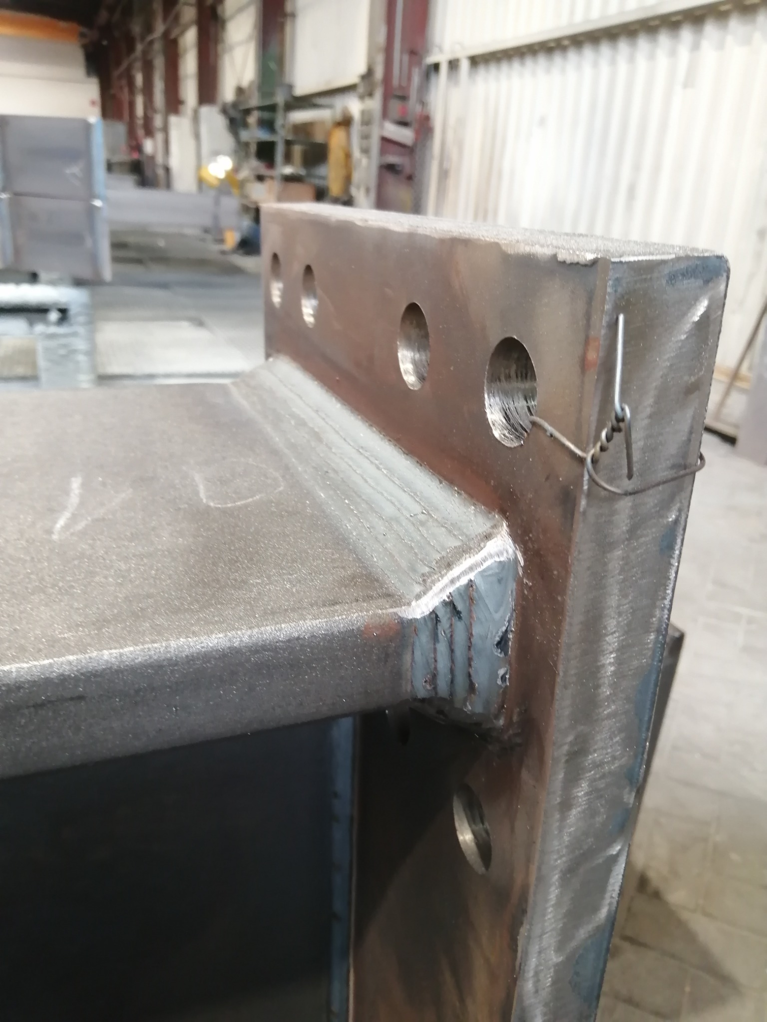 Metalowa konstrukcja spawana, z widocznymi śladami spawania i otworami, zabezpieczona drutem, w hali produkcyjnej.