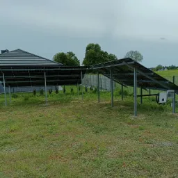 Naziemna instalacja paneli słonecznych przy domu jednorodzinnym z widocznym inwerterem i ogrodzeniem z siatki.