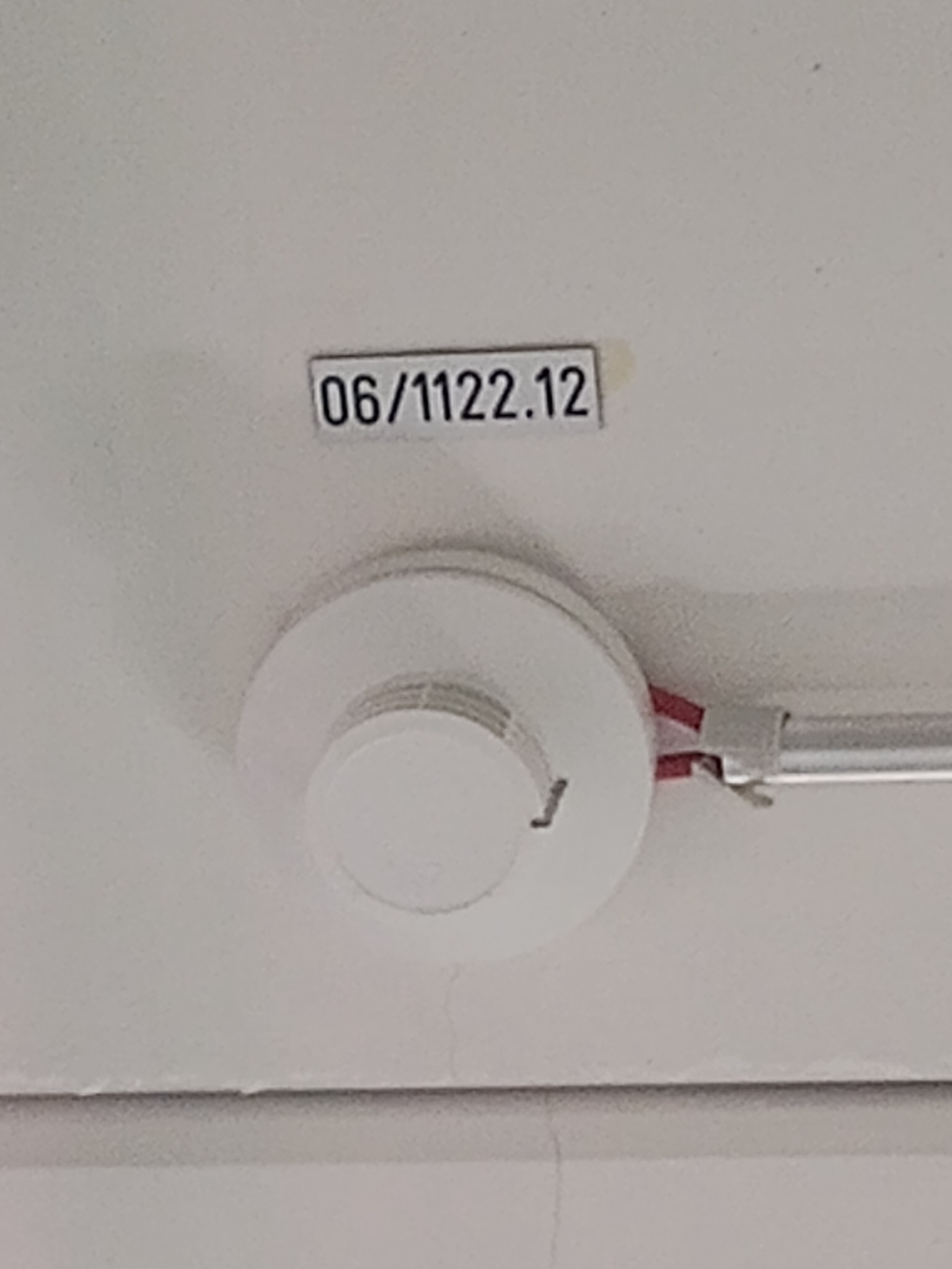 Biały czujnik dymu zamontowany na suficie, z widoczną etykietą '06/1122.12' powyżej i fragmentem instalacji elektrycznej z czerwonymi elementami.