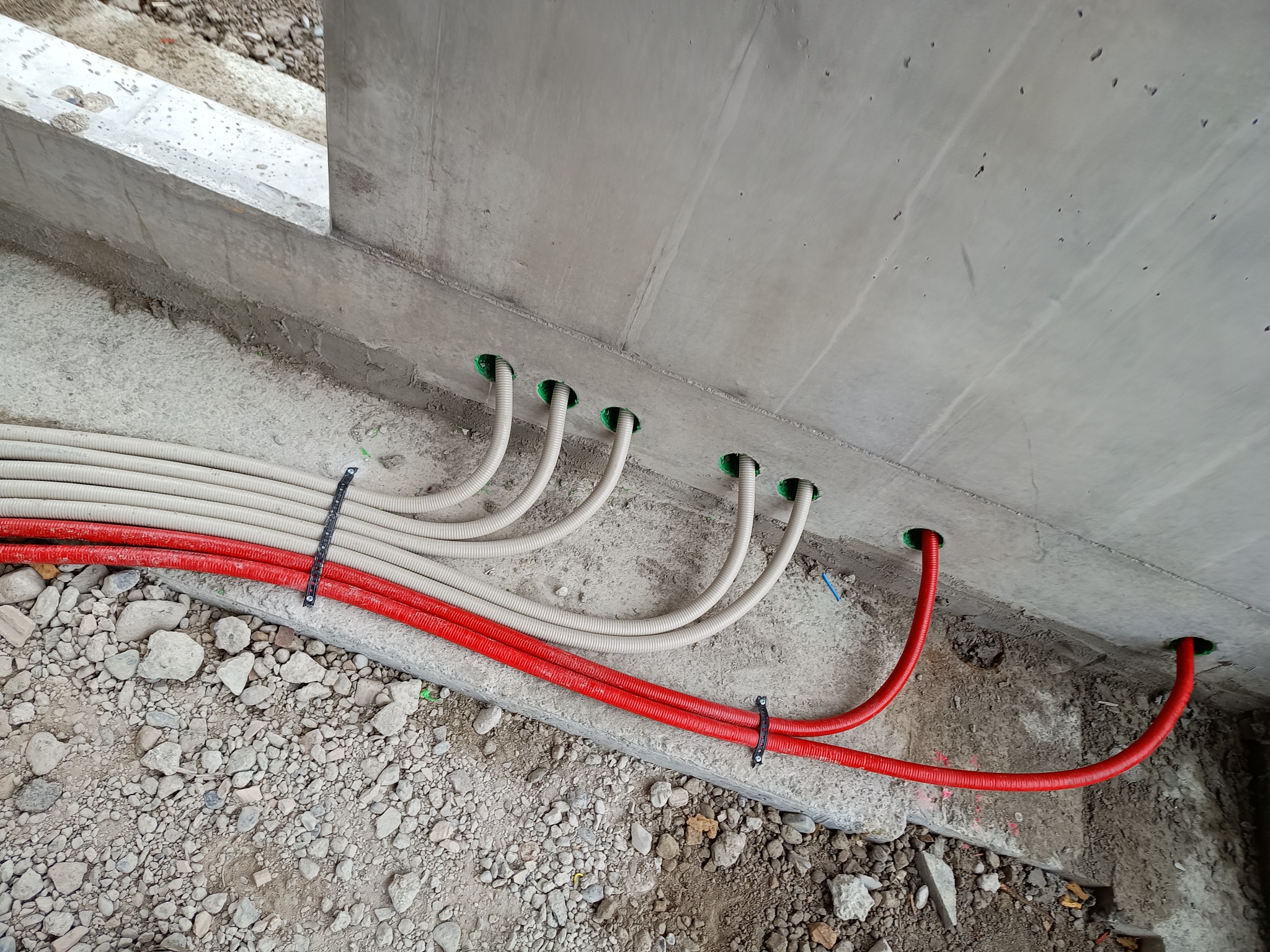 Rury instalacyjne w kolorze białym i czerwonym wchodzące w otwory w betonowej ścianie, zabezpieczone czarną opaską zaciskową, widoczne gruz i kamienie na podłożu.