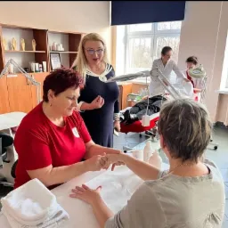 Kurs manicure: Instruktor w czerwonej koszulce demonstruje technikę manicure na dłoni kursantki, nadzorowane przez nauczycielkę w granatowej sukience, w tle inni kursanci ćwiczą na modelach.