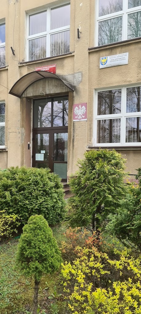 Wejście do budynku z oznaczeniami urzędowymi, otoczone zielenią.