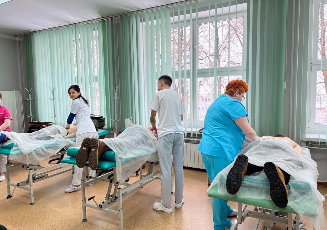 Sala szkoleniowa z trzema stanowiskami do masażu; kursanci w trakcie praktyki masażu na leżących osobach, widok z perspektywy kursanta; jasne wnętrze z zielonymi żaluzjami.