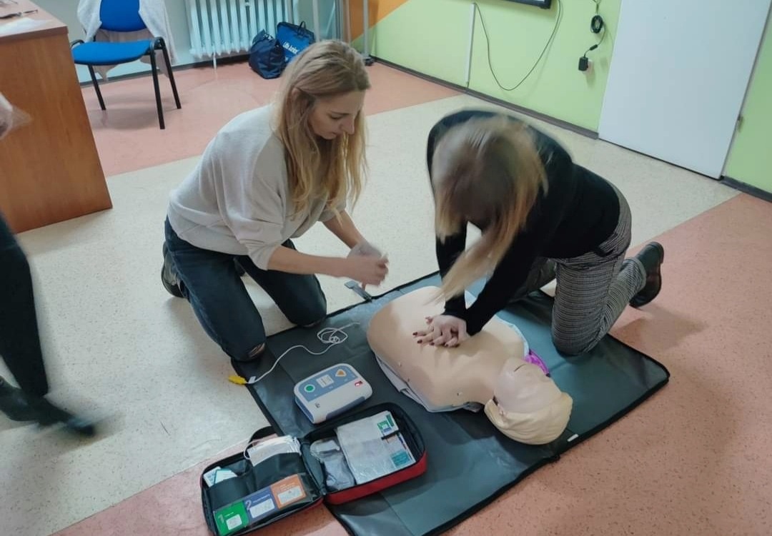 Dwie kobiety ćwiczą resuscytację krążeniowo-oddechową na manekinie, obok defibrylator i apteczka pierwszej pomocy.