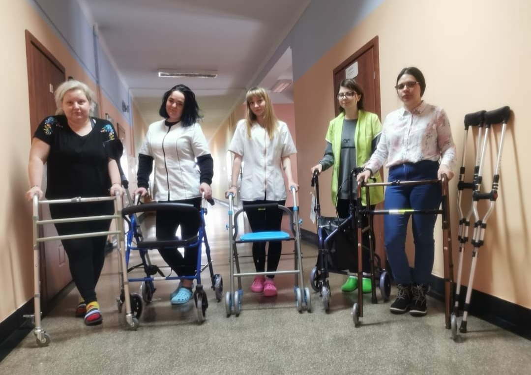 Pięć kobiet w korytarzu, trzymających różne rodzaje chodzików i kul ortopedycznych, prawdopodobnie podczas szkolenia z rehabilitacji.