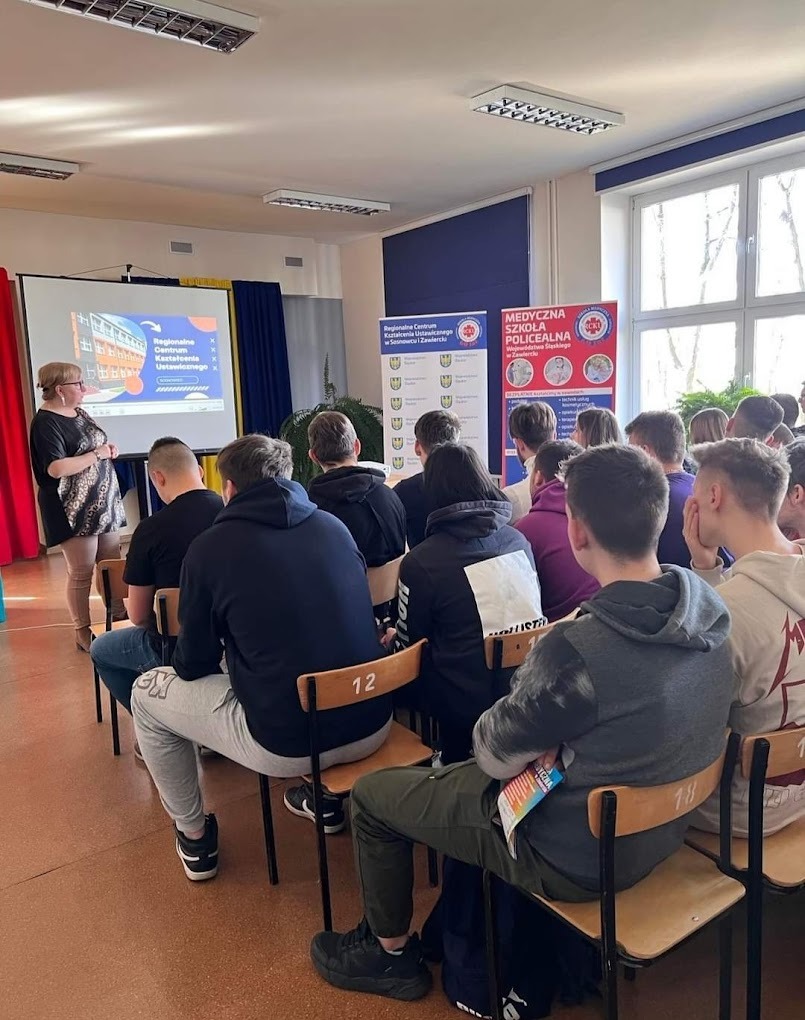 Sala lekcyjna: kobieta w czarnej sukience prowadzi prezentację na rzutniku dla grupy uczniów siedzących na krzesłach z numerami. W tle widoczne plakaty informacyjne.