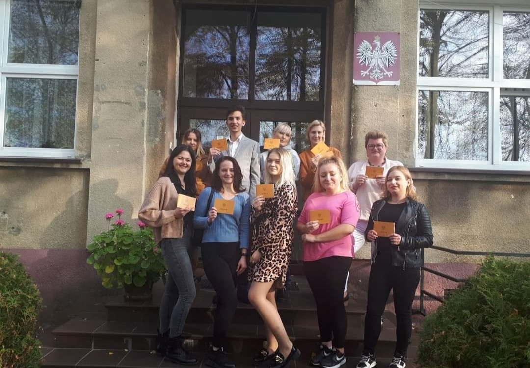 Grupa absolwentów pozuje z dyplomami na schodach budynku z godłem Polski.