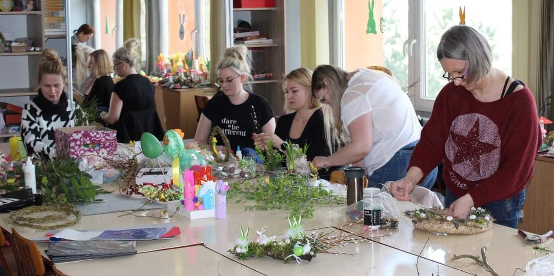Grupa kobiet uczestniczy w warsztatach rękodzielniczych, tworząc dekoracje wielkanocne z gałązek, kwiatów i ozdobnych jajek.