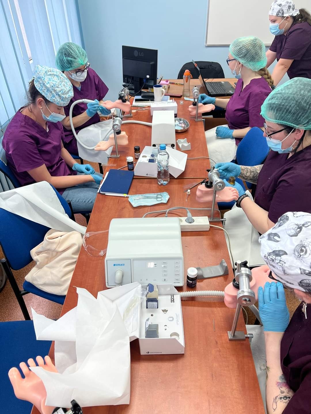 Grupa osób w odzieży ochronnej ćwiczy zabiegi podologiczne na modelach stóp przy użyciu specjalistycznego sprzętu i narzędzi.