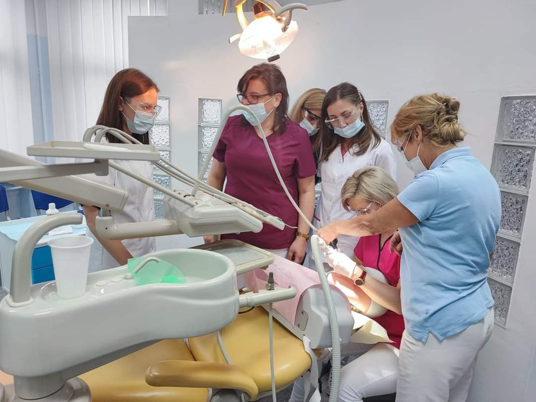 Grupa osób w maseczkach obserwuje dentystkę w różowej bluzie podczas pracy przy pacjencie na żółtym fotelu dentystycznym, w tle widoczne szklane pustaki.