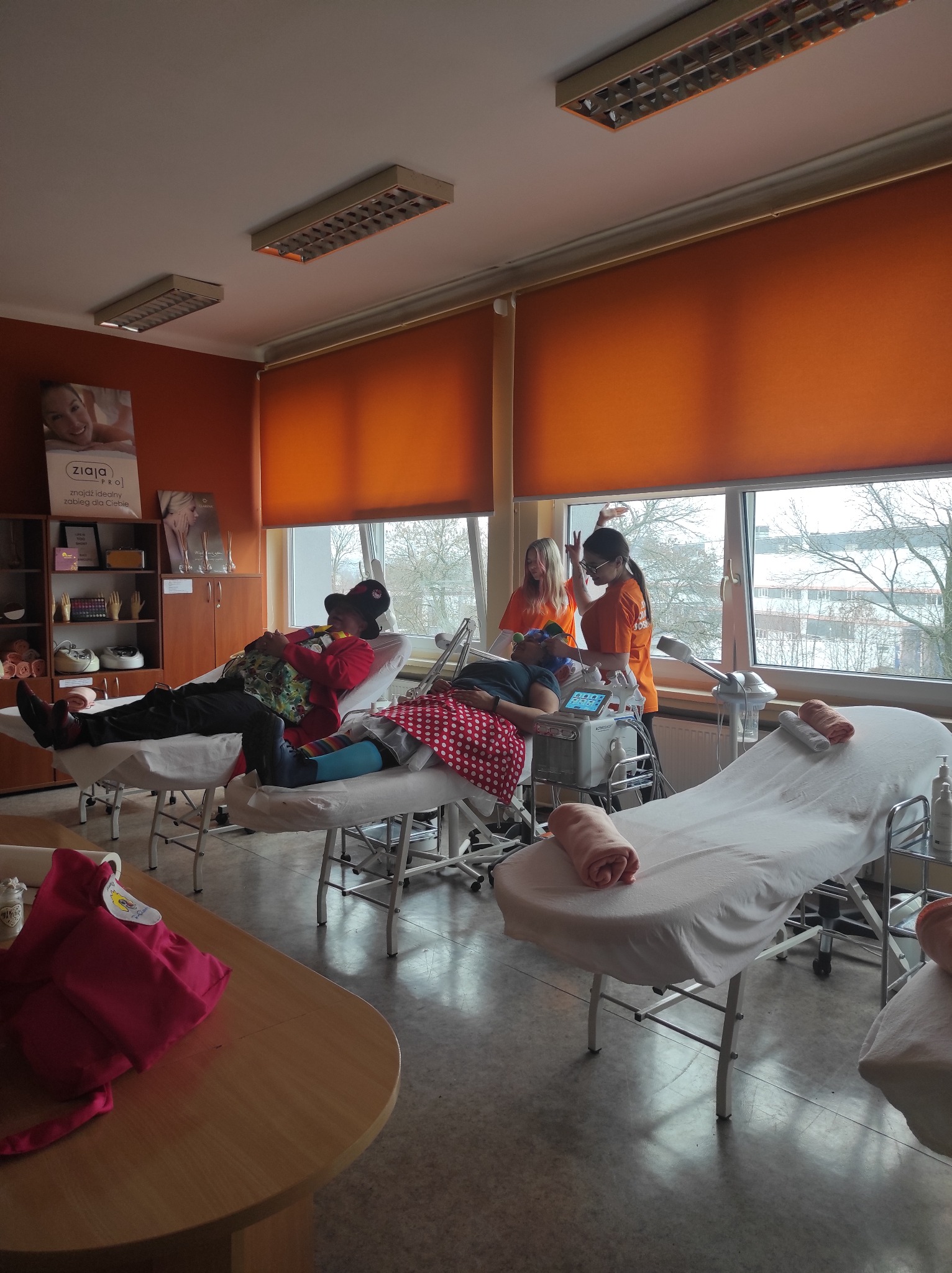 Szkolenie z zakresu kosmetyki: kursanci leżą na leżankach podczas zabiegu, instruktorki w pomarańczowych koszulkach obsługują urządzenie kosmetyczne, w tle okno i regał z produktami Ziaja Pro.