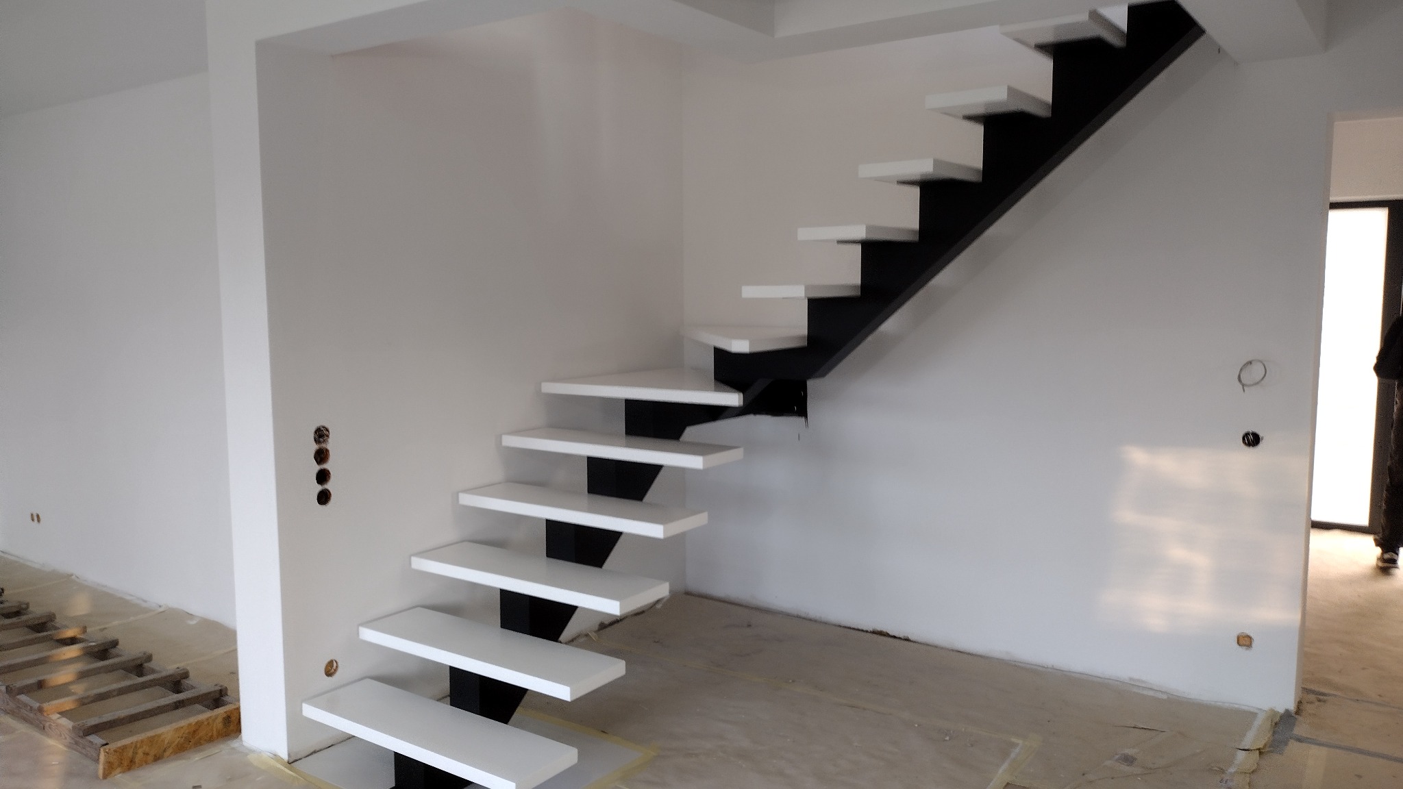 Wnętrze w trakcie wykańczania z nowoczesnymi schodami wspornikowymi o białych stopniach i czarnej konstrukcji nośnej, widoczne przygotowania do montażu podłogi.