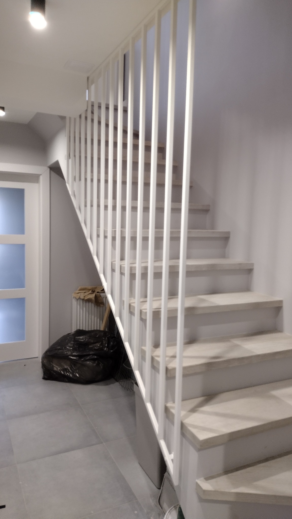 Nowoczesne schody z białymi, pionowymi poręczami, widoczne z dołu, z workiem na śmieci i grzejnikiem w tle.