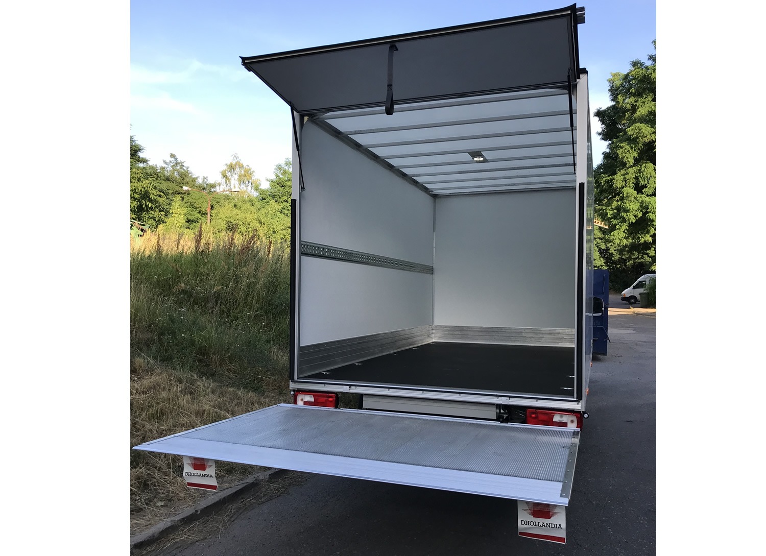 Otwarta przestrzeń ładunkowa busa z podnośnikiem, widok z tyłu na puste wnętrze i otwartą klapę, na zewnątrz zieleń.