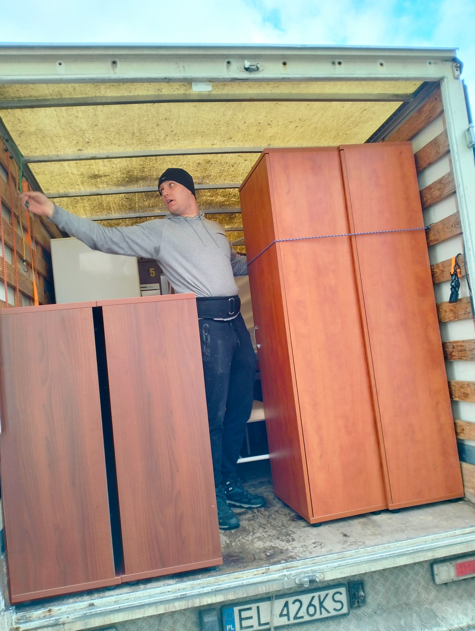 Mężczyzna w szarej bluzie i czapce, z pasem lędźwiowym, zabezpiecza meble wewnątrz przestrzeni ładunkowej samochodu dostawczego o żółtym dachu, widoczne szafy z ciemnego drewna, liny mocujące...