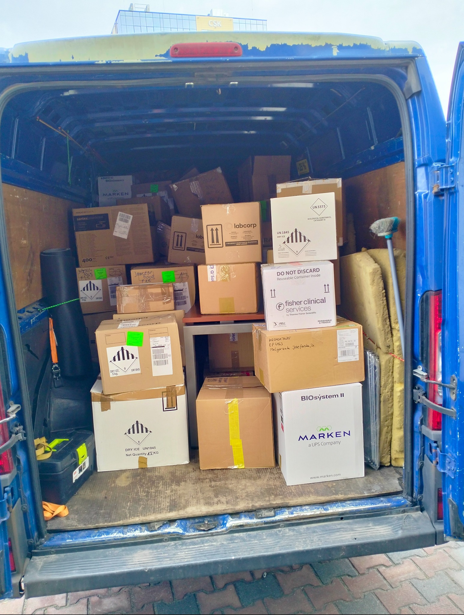 Wnętrze niebieskiego busa wypełnione kartonami różnych rozmiarów, niektóre z oznaczeniami 'Dry Ice' i 'Biological Substance', sugerujące transport materiałów laboratoryjnych.