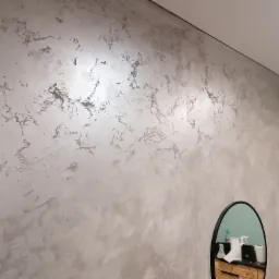 Ściana z dekoracyjną, srebrzystą fakturą, odbijająca się w owalnym lustrze z czarną ramą, na tle białego sufitu.