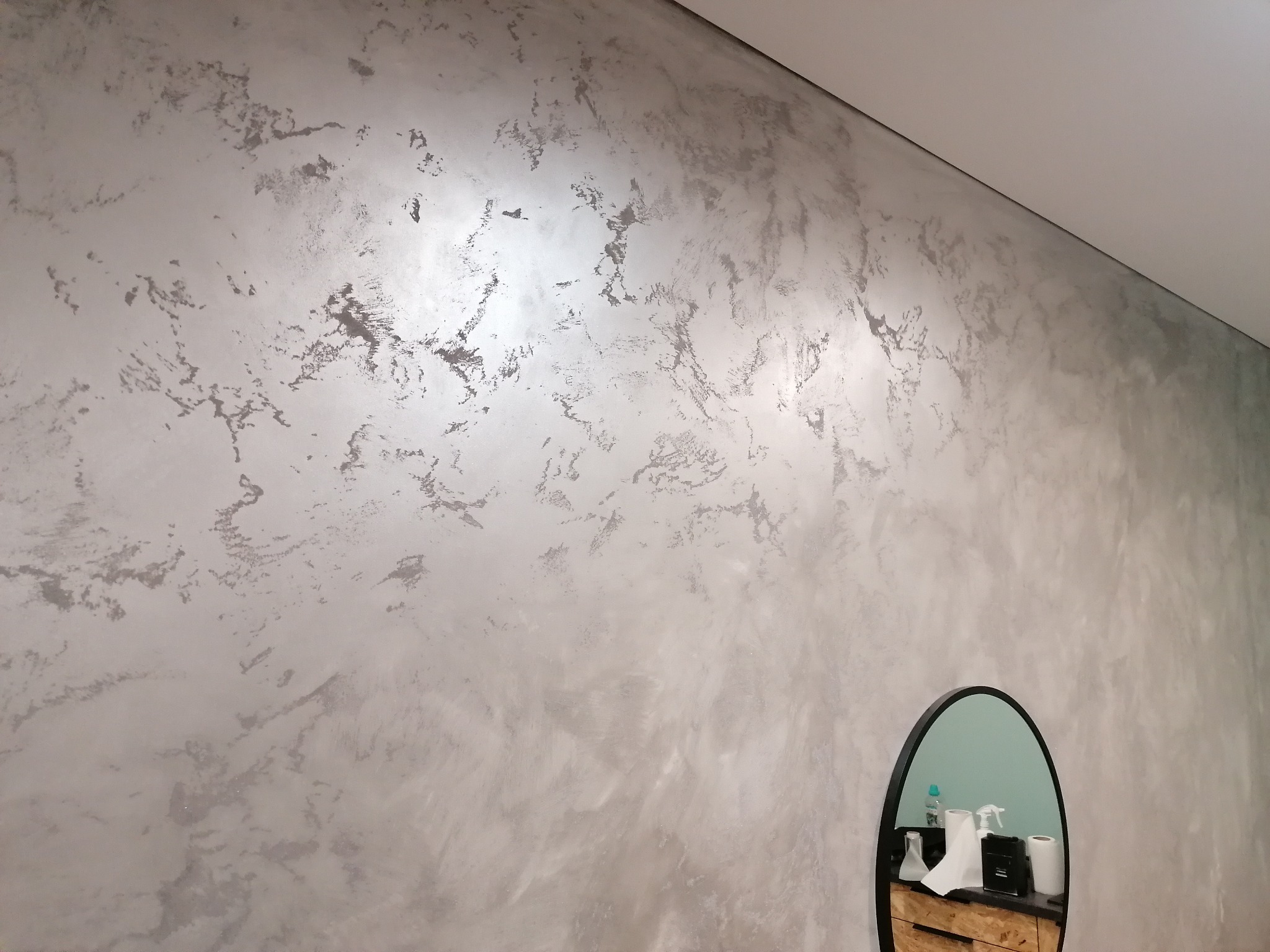 Ściana z dekoracyjną, srebrzystą fakturą, odbijająca się w owalnym lustrze z czarną ramą, na tle białego sufitu.