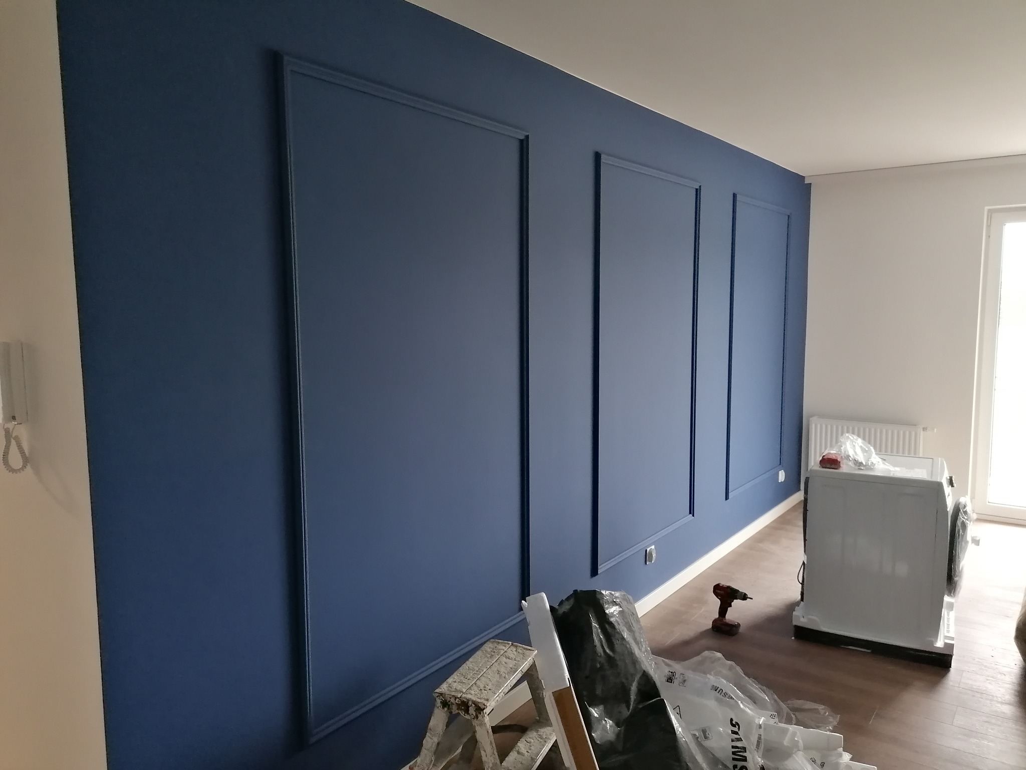 Pomieszczenie w trakcie malowania na granatowo z dekoracyjnymi panelami ściennymi, widoczna drabina, wiertarka akumulatorowa i pralka w folii ochronnej.